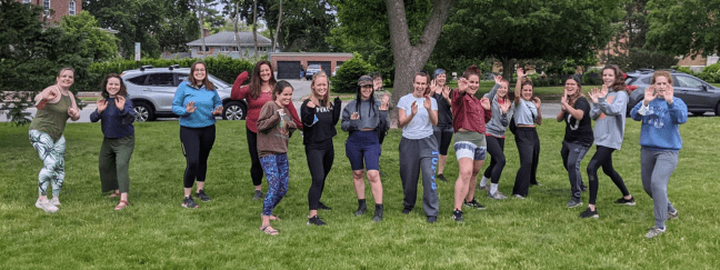 A large group of participants stand in a grassy park in a ready stance with their hands raised, practicing self-defense techniques together.