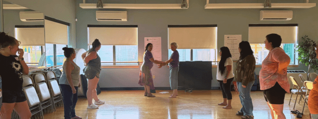 A group of participants stand in a circle in a studio space while two people demonstrate a hands-on exercise in the center.