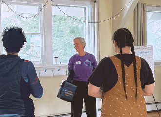 An instructor in a purple Prevention. Action. Change. shirt leads a group of participants standing in a circle during a self-defense training in a bright room.