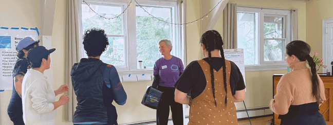 An instructor in a purple Prevention. Action. Change. shirt leads a group of participants standing in a circle during a self-defense training in a bright room.