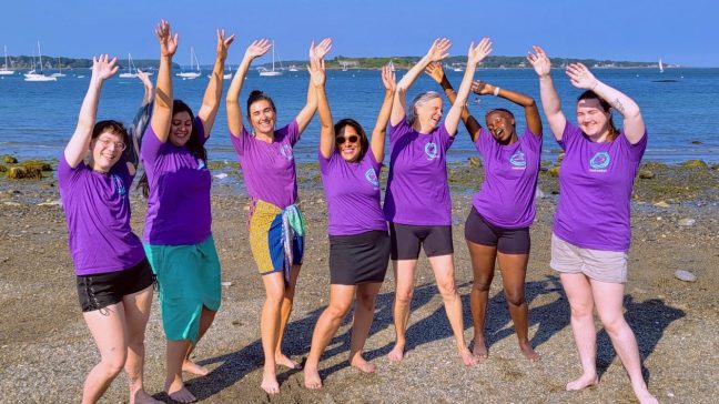 Seven Empowerment Self-Defense instructors wearing matching purple instructor T-shirts stand barefoot on a sandy beach with their arms raised and smiling toward the camera. Calm blue water, sailboats, and a clear blue sky stretch across the background, creating a joyful, celebratory scene.